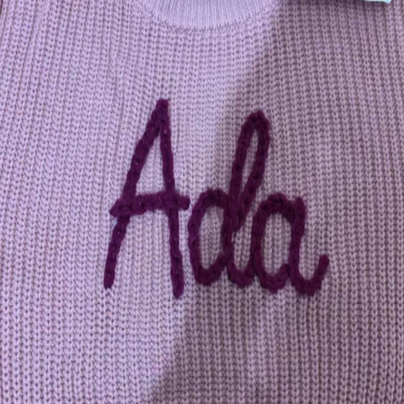 Nahaufnahme des handbestickten Namens Ada in dickem Garn auf einem lila Pullover
